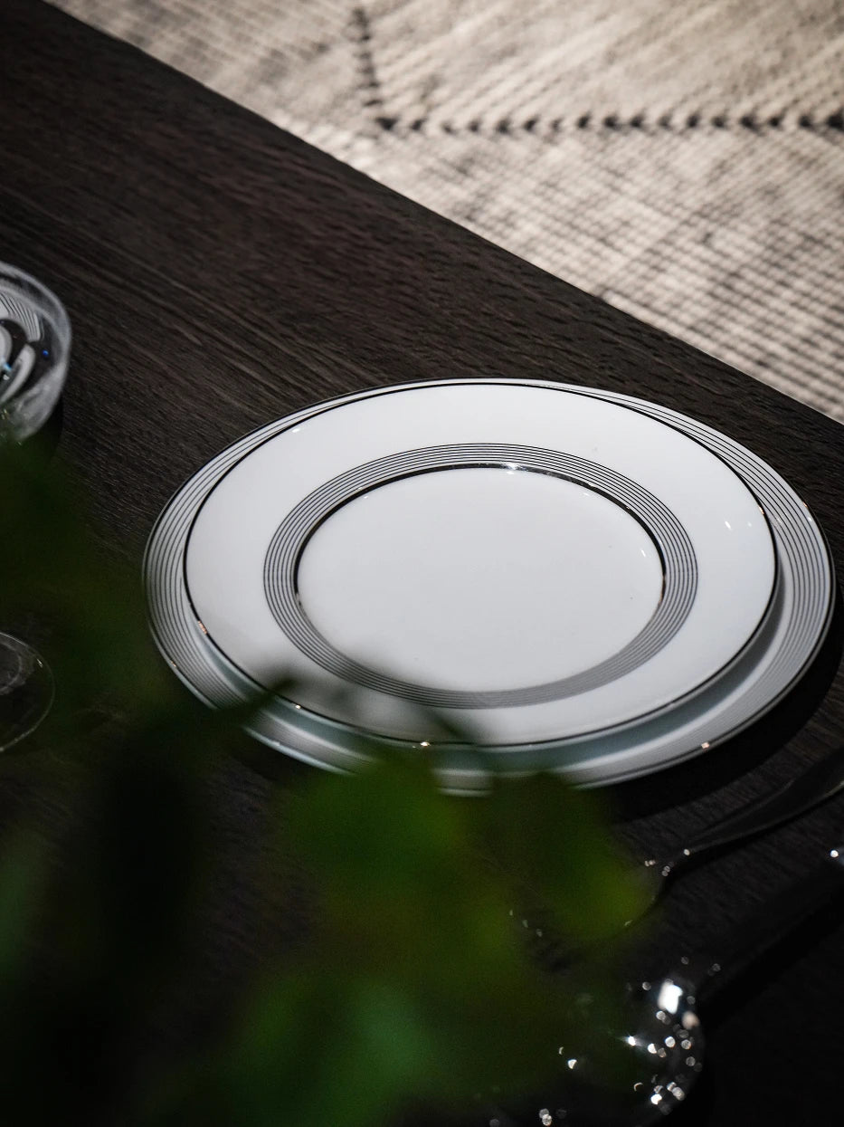 Minimal close-up shot of ERYA medium black-silver plate on dark wood tabletop, soft blurred green foliage as foreground decoration
