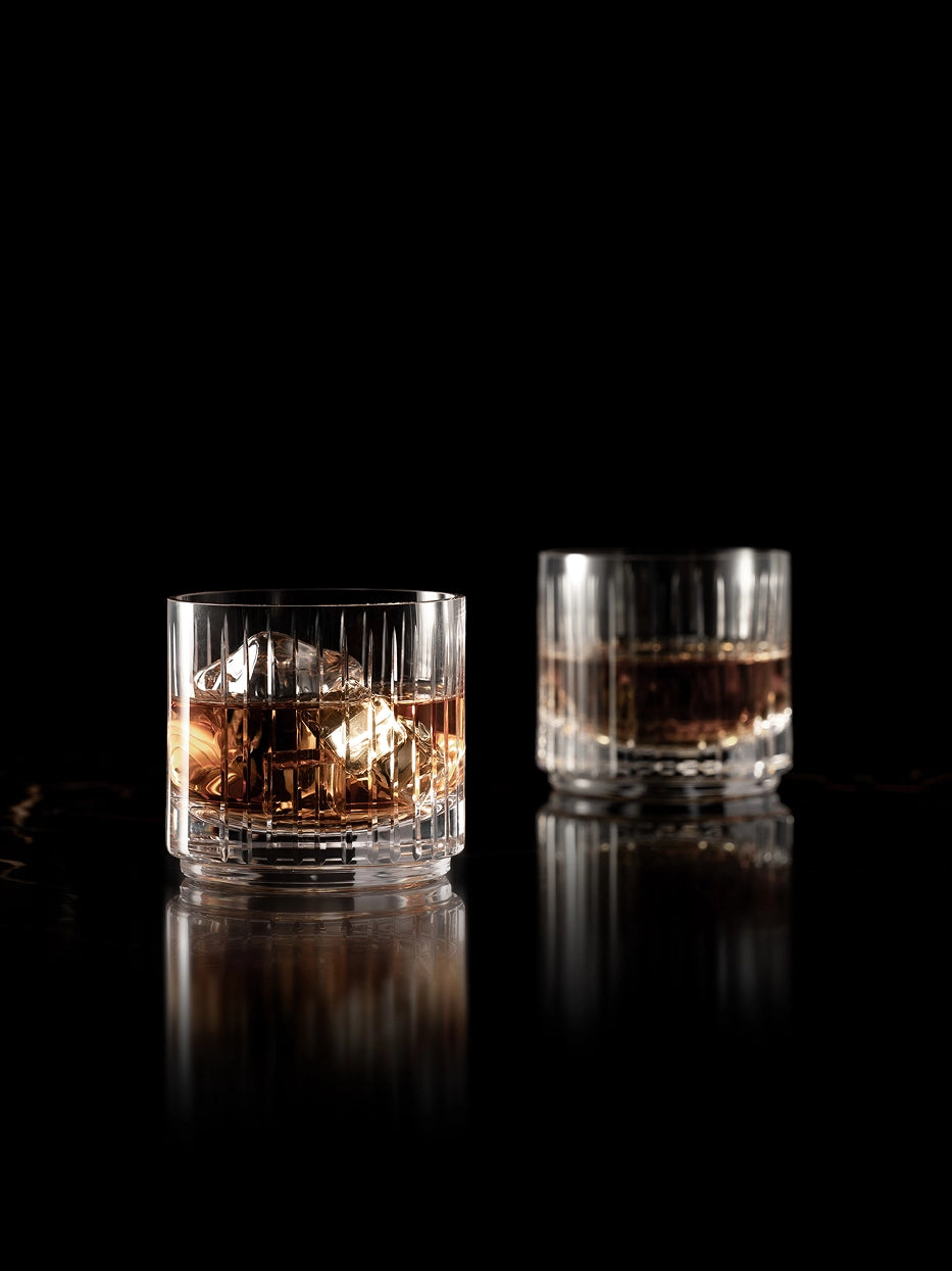 Luxury dark studio shot of two FILO fluted crystal whiskey tumblers filled with amber whiskey & large ice cubes, reflective black glossy tabletop with mirror reflection