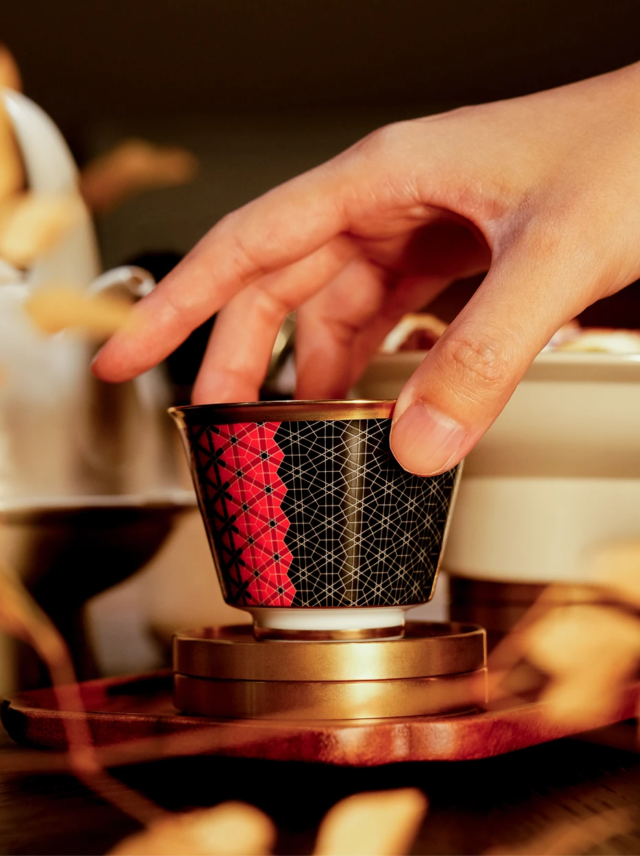 Human hand close-up shot holding GUOYUE Black & Red ceramic cup, blurred tea foliage and dessert as foreground to create layered warm texture