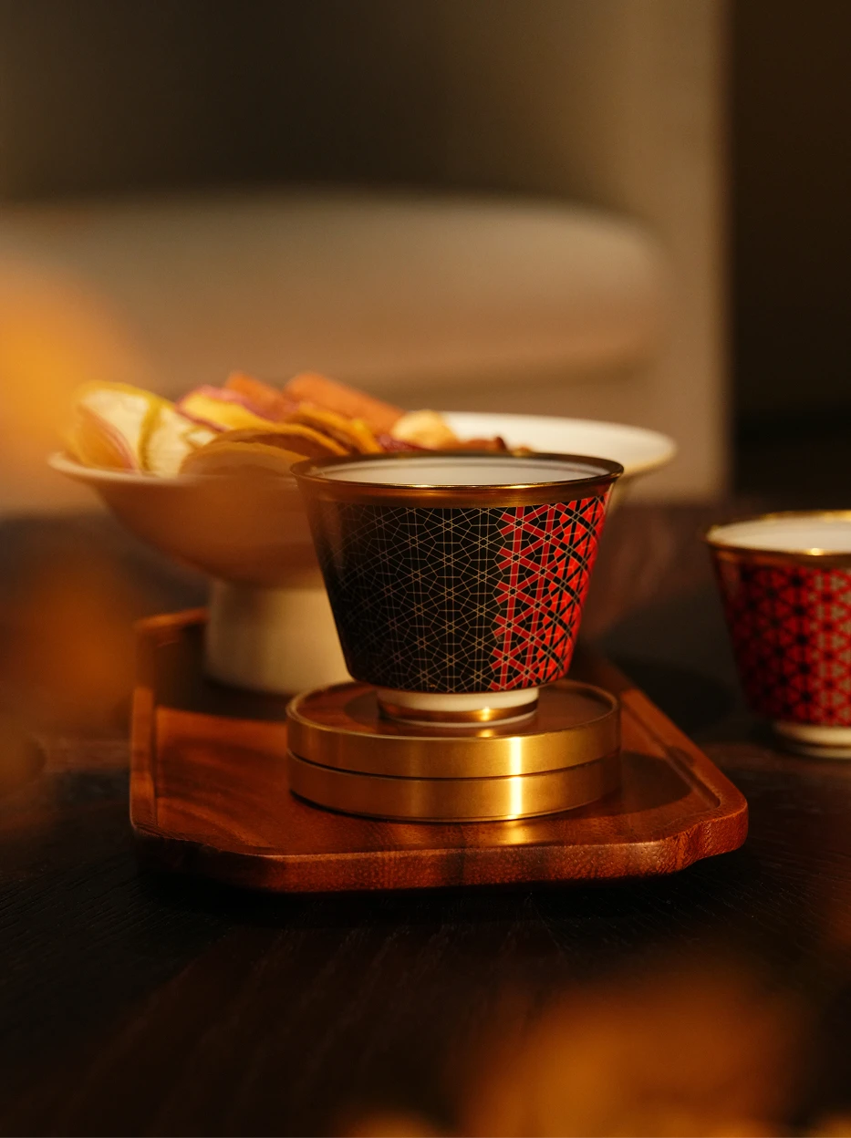 Warm afternoon tea lifestyle shot of GUOYUE Black & Red gold-rim ceramic cup placed on walnut wood tray, double-layer metal coaster and dessert plate as decor, dim warm indoor light
