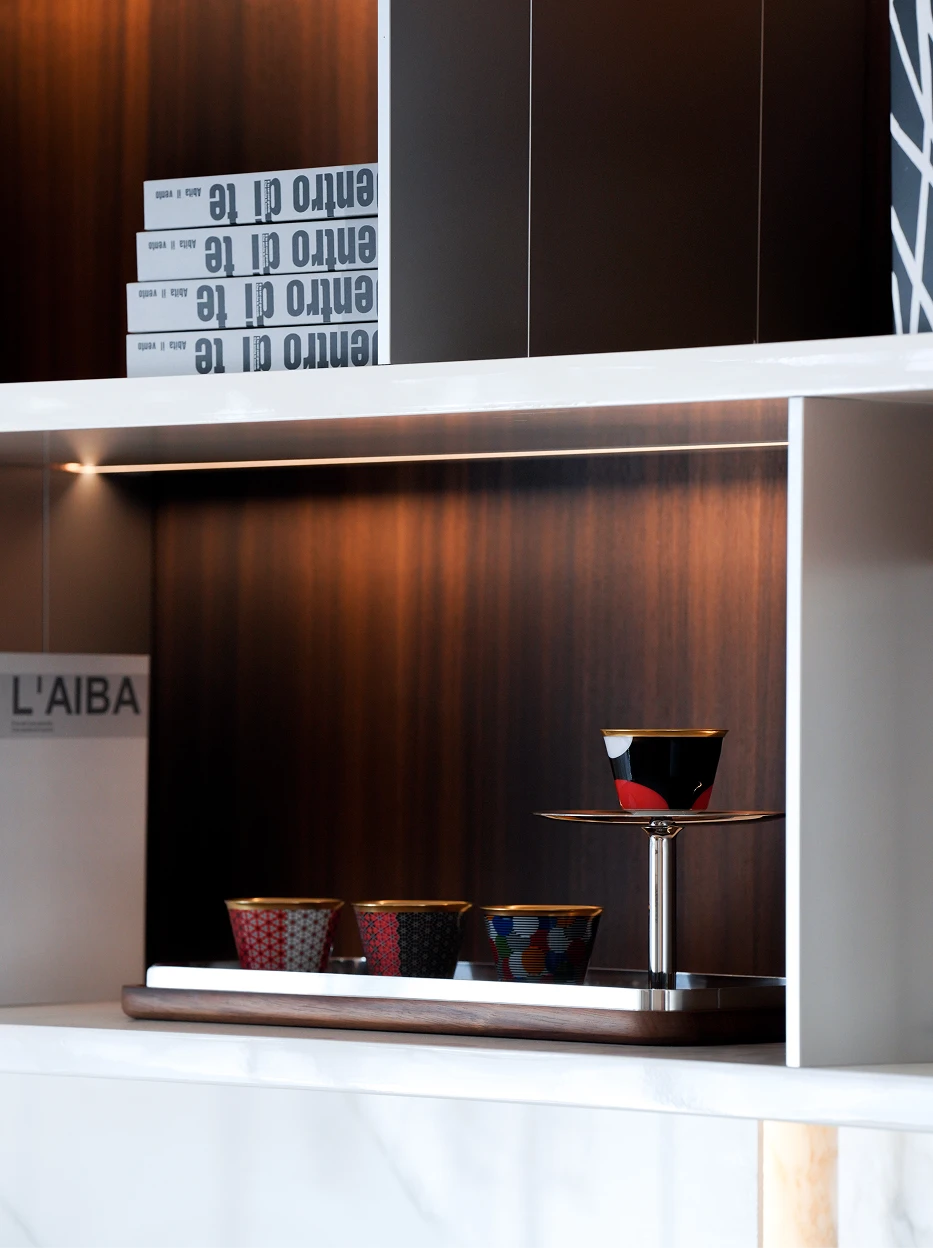 Modern luxury interior shot of GUOYUE Sunrise gold-rim cup displayed on two-tier metal tray inside white & dark wood open cabinet, matched with other GUOYUE colorway cups and stacked art books