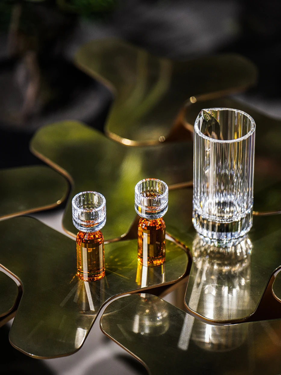Dark luxury close-up of HAOYING amber base crystal baijiu set laid on irregular reflective metal coffee table, soft ambient shadow highlighting two-tone glass texture