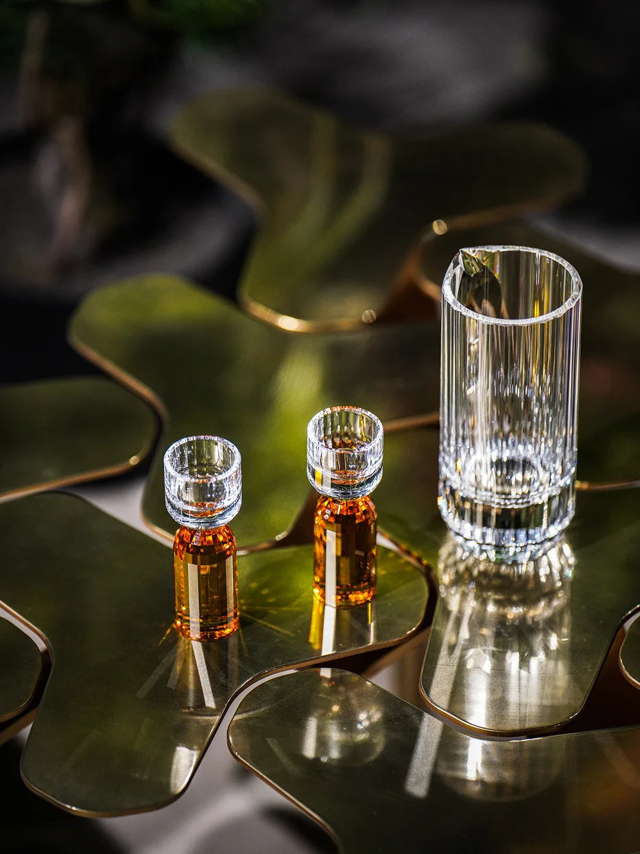 Dark luxury close-up of HAOYING amber base crystal baijiu set laid on irregular reflective metal coffee table, soft ambient shadow highlighting two-tone glass texture