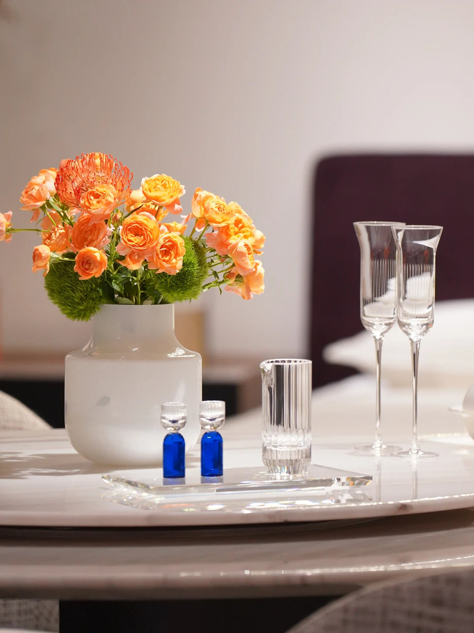 Warm cozy living room shot of HAOYING blue base crystal baijiu set on clear rectangular tray, matched with orange roses ceramic vase on white marble round table
