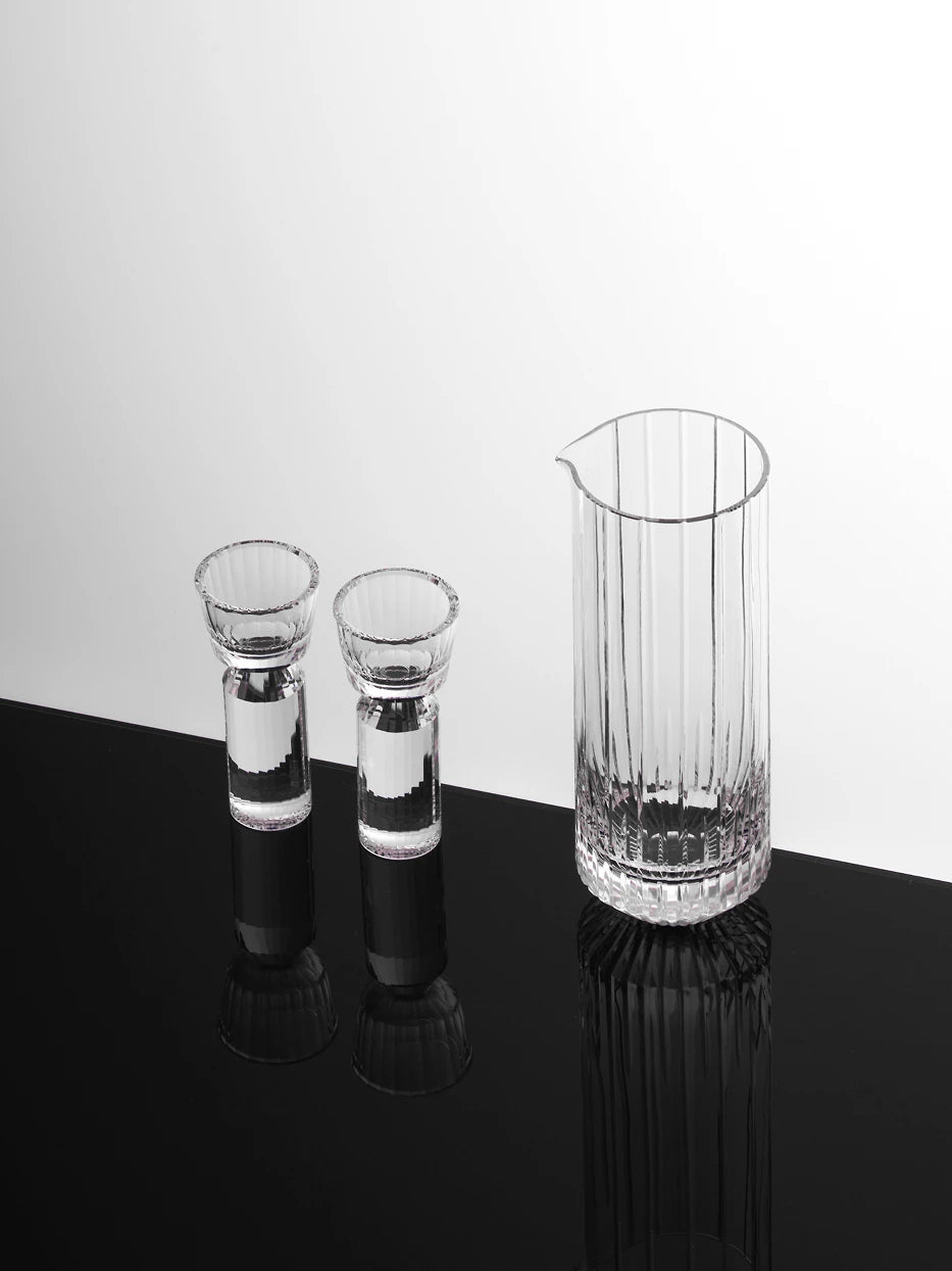 Minimalist studio shot of partial HUAZUN clear crystal baijiu set, 2 faceted shot glasses matched with 1 vertical cut pitcher, reflective black tabletop with complete mirror reflection
