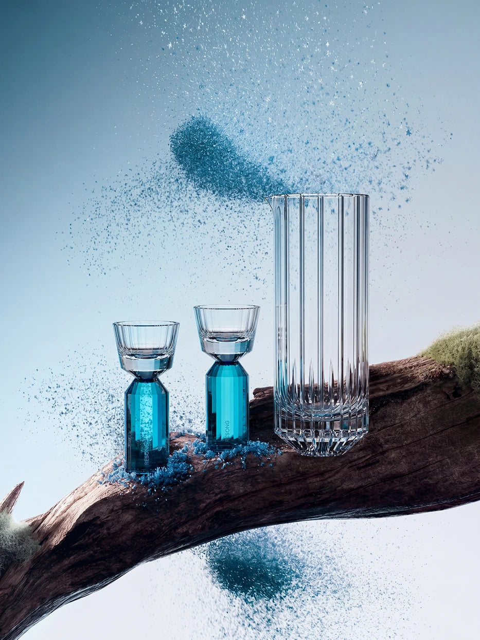 Artistic creative lifestyle shot of full HUAZUN lake blue crystal baijiu set laid on natural dry wood branch, decorated with fine blue sand particles, gradient light blue sky background