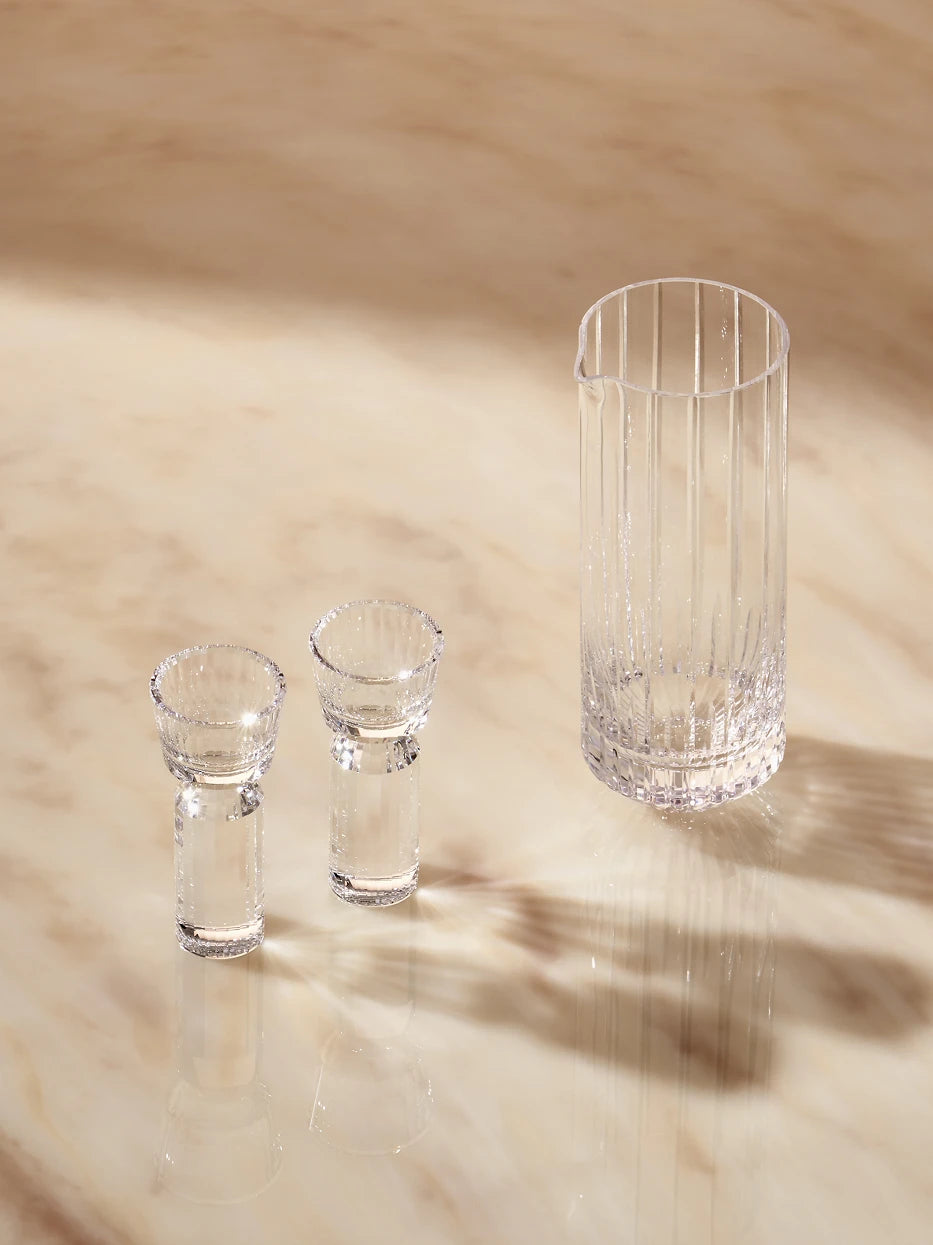 Cozy elegant lifestyle shot of HUAZUN clear crystal baijiu set laid on light polished marble table, soft warm natural ambient light creating striped glass shadows