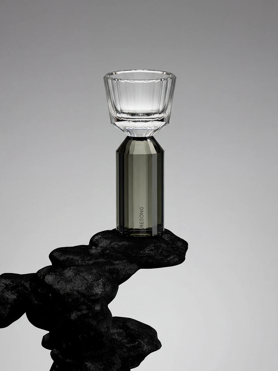 Artistic minimalist close-up of single HUAZUN smoke gray faceted shot glass framed by rough black natural rock blocks, pure light gray negative space background