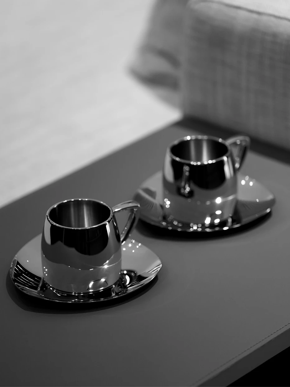Black and white atmospheric still life of two SHEDI large mirror steel cup & saucer sets on solid grey matte tabletop, soft reflective bokeh light