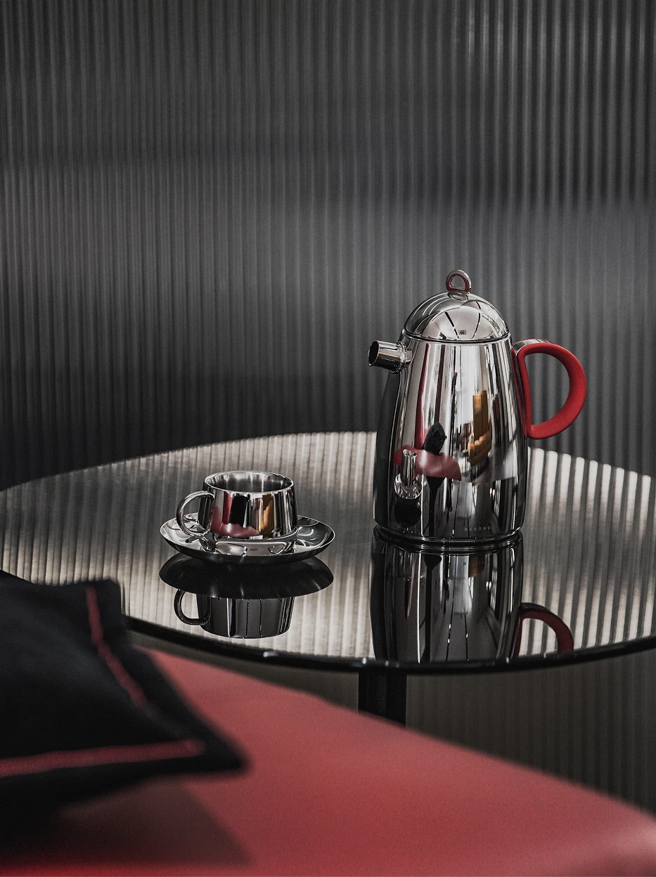 Atmospheric close-up shot of large SHEDI red handle teapot and matching metal cup saucer set on reflective round table against vertical striped wall
