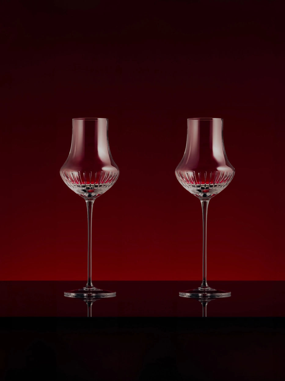 Luxury studio display of two TRIUMPH fluted crystal wine stem glasses with red wine, reflective black glossy countertop against solid deep burgundy backdrop