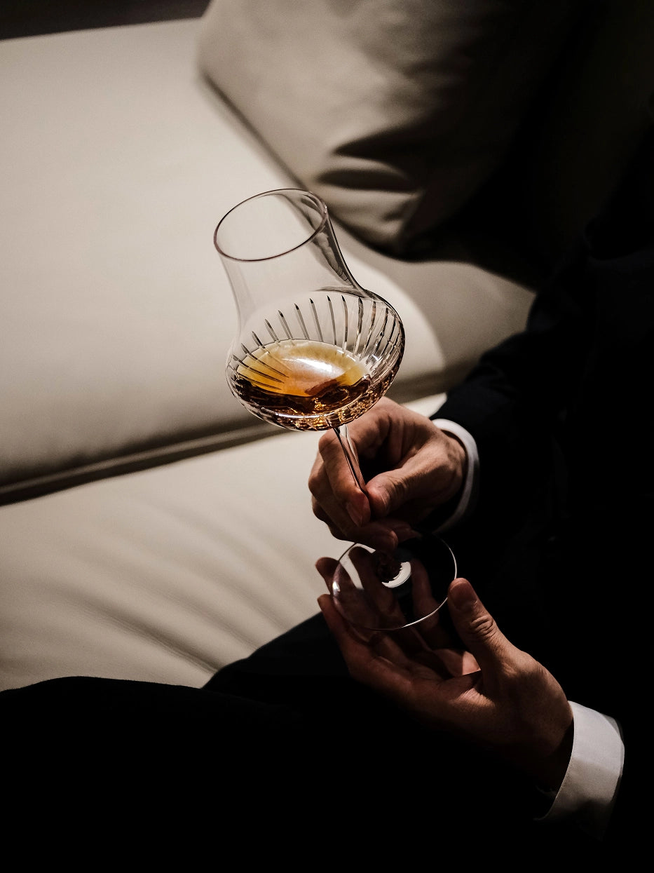 Cozy sophisticated lifestyle shot of hands holding TRIUMPH carved crystal wine glass with amber liquor beside soft light fabric sofa, dim warm indoor ambient light