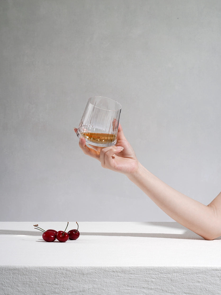 Soft minimalist lifestyle shot of hand holding TRIUMPH fluted crystal whiskey tumbler filled with amber liquor, fresh cherries on linen white table surface