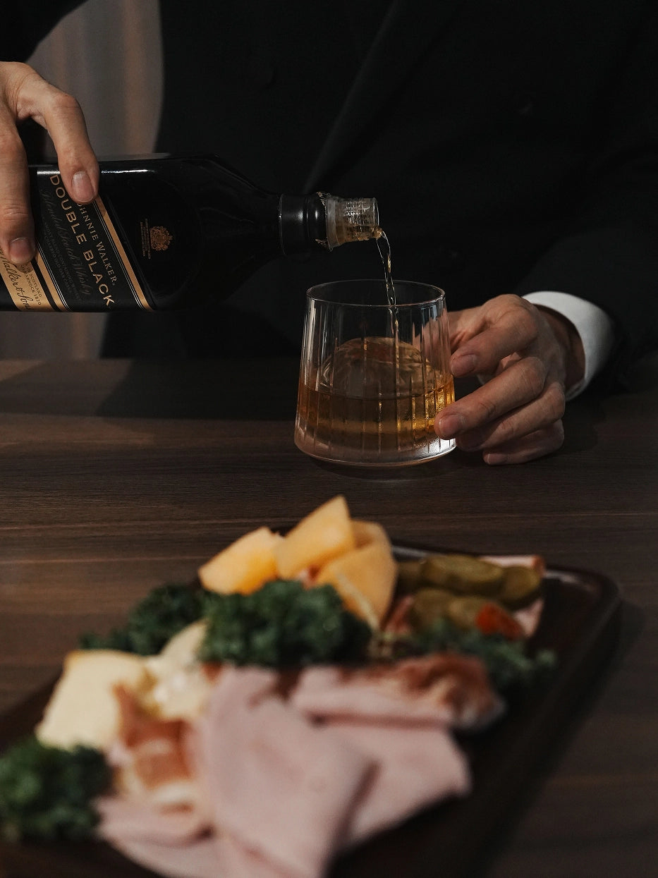 Sophisticated bar lifestyle photo pouring scotch into TRIUMPH vertical fluted crystal tumbler, paired with charcuterie cold cuts platter on dark wooden dining table