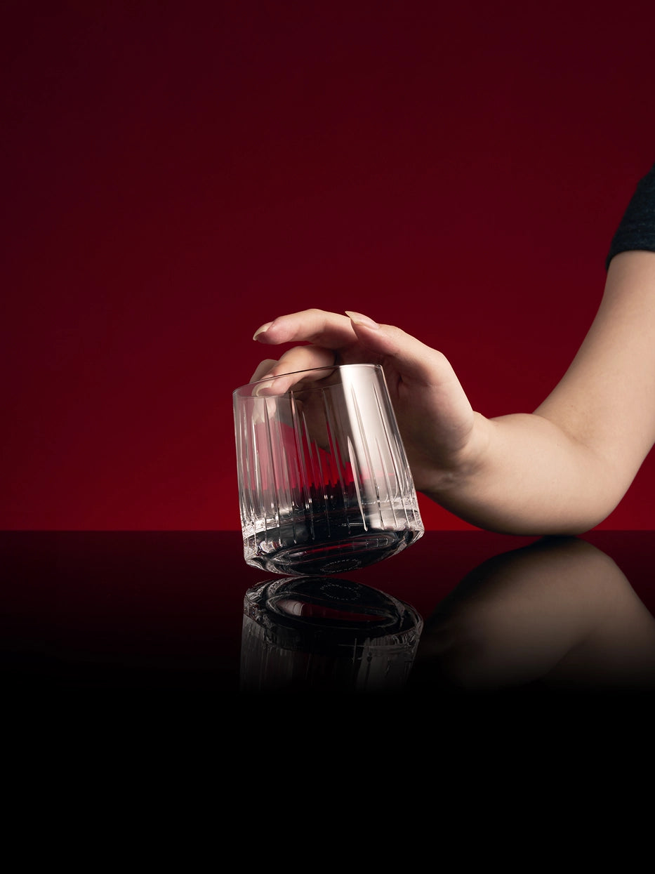 Luxury dark studio shot, hand gripping empty TRIUMPH carved crystal whiskey glass, reflective black tabletop with mirror reflection against solid burgundy background
