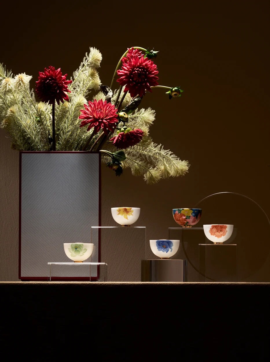 Light luxury showroom display shot of five underglaze multicolour tea cups on layered acrylic stands, matched with red dahlia floral arrangement as background decor