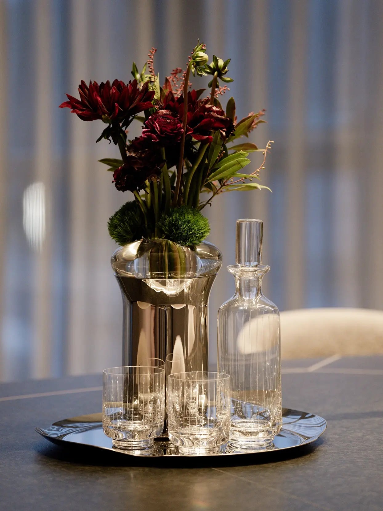 Elegant dining interior photo of XUYU etched crystal whiskey decanter & tumbler set on glossy metal tray, styled with metallic flower vase filled with burgundy dahlias on dark slate tabletop