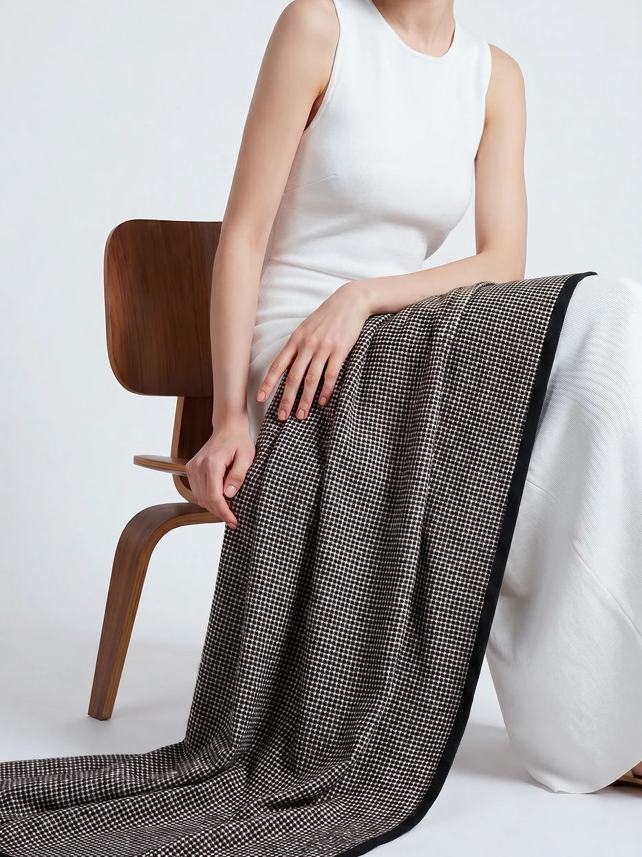 Minimalist full-body portrait shot of model sitting on wooden chair draped with LUODI retro check cashmere throw, fully displays overall blanket silhouette, full-width tiny check layout and black contrast edge design, dual function as decorative home couch cover and wearable shawl for indoor & outdoor use