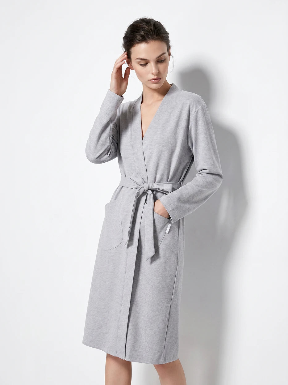 Pure white background full-body side shot of model with hands in pockets wearing size M MAYA light gray stretch bathrobe, showing loose dropped shoulder moderate long silhouette, soft draping light heather gray texture, highlighting delicate skin-friendly stretch silk cotton wearing experience