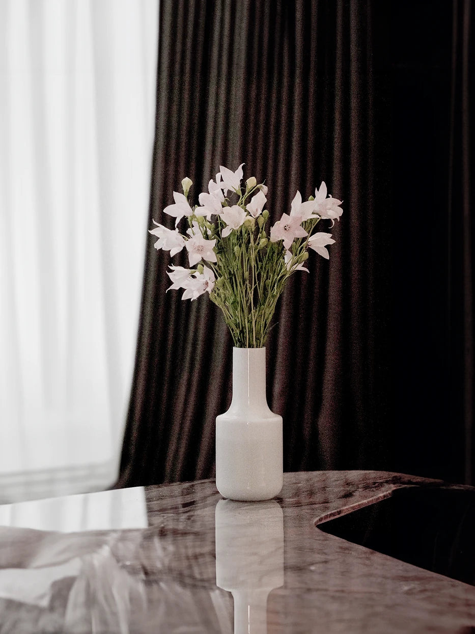 Soft minimalist home furnishing shot of white minimalist vase A full of light pink bellflowers on glossy marble side table beside dark brown striped curtain