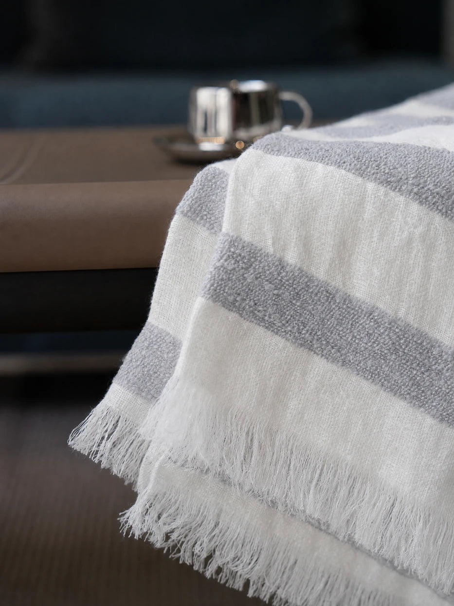 Extreme close-up texture shot of MUJIE linen wool blend throw blanket, showing bumpy woven grey-white stripe texture and loose handmade tassel fringe detail, visible breathable linen weave and soft wool mixed touch, neatly cut natural fringed edges, displays premium fabric craft and tactile texture for home textile product display