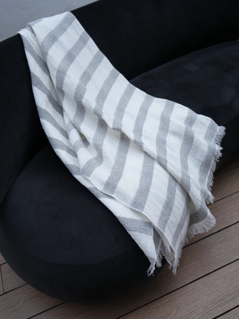 Minimalist interior still shot of MUJIE striped linen throw draped over curved black accent armchair on solid wood flooring, soft contrast between light grey-white textile and dark furniture, lightweight breathable fabric without bulky texture, great decor for entryway lounge, reading nook, hotel rest corner, modern dark tone interior matching