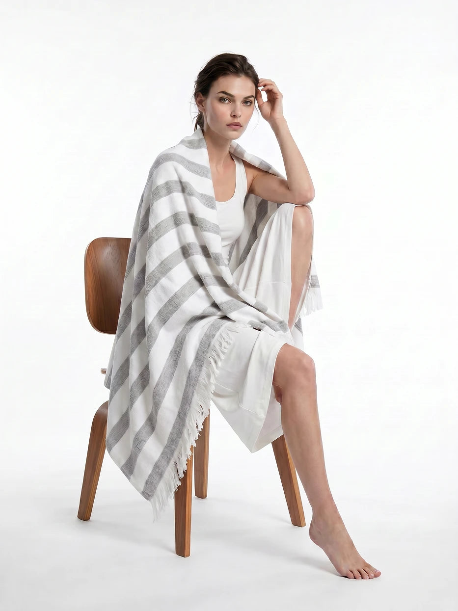 Minimalist full portrait shot of model seated on solid wood chair wrapped with MUJIE gray linen striped throw, fully displays overall blanket silhouette, complete stripe layout and bottom fringe design, dual function as decorative home couch cover and wearable shawl for indoor & outdoor use