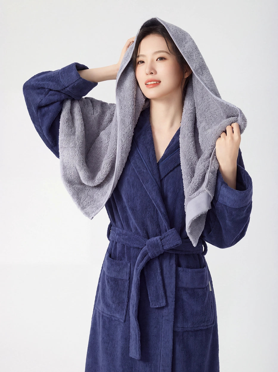 Woman wearing RESONG navy blue hooded terry cloth bathrobe, holding a grey cotton bath towel over her head wrapping wet hair, luxury bath towel and bathrobe gift set, white studio background
