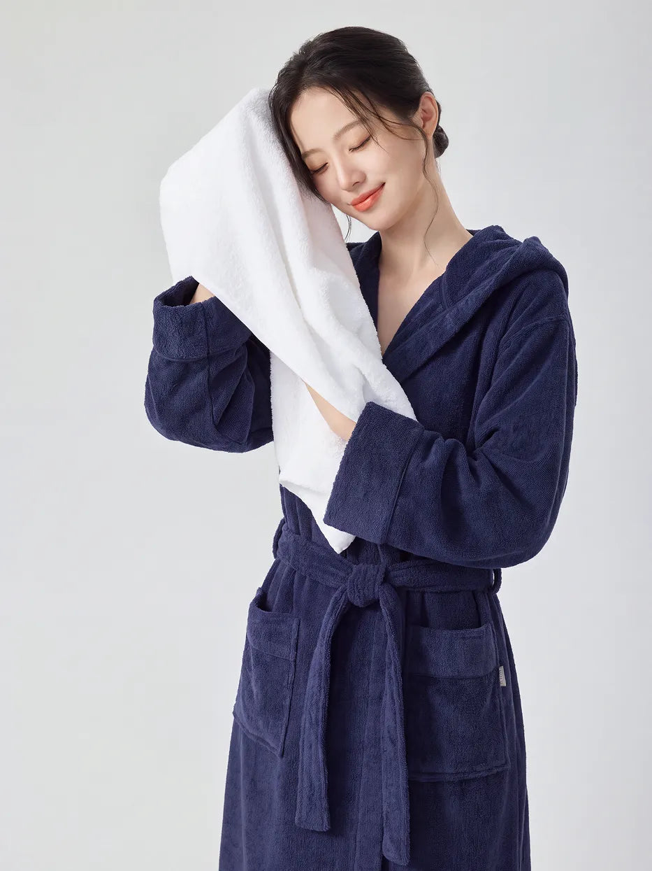 Woman wearing RESONG navy blue hooded terry cloth bathrobe, eyes closed gently pressing a white cotton bath towel against her cheek to dry face, luxury bath towel and bathrobe gift set, white studio background
