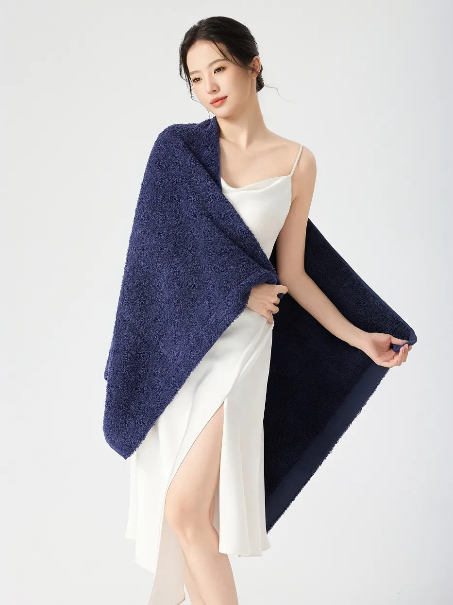 Woman standing draped in RESONG navy blue cotton terry bath towel over one shoulder, gently holding towel corner showing fluffy absorbent texture, white slip dress with side slit, white studio background
