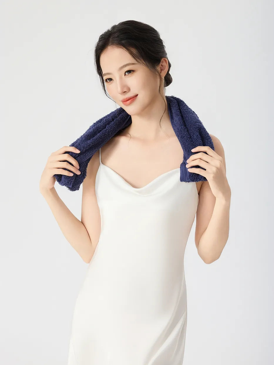 Woman in white slip dress smiling, RESONG navy blue cotton terry face towel draped around neck like a scarf, holding both towel ends, soft fluffy face towel on model, white studio background
