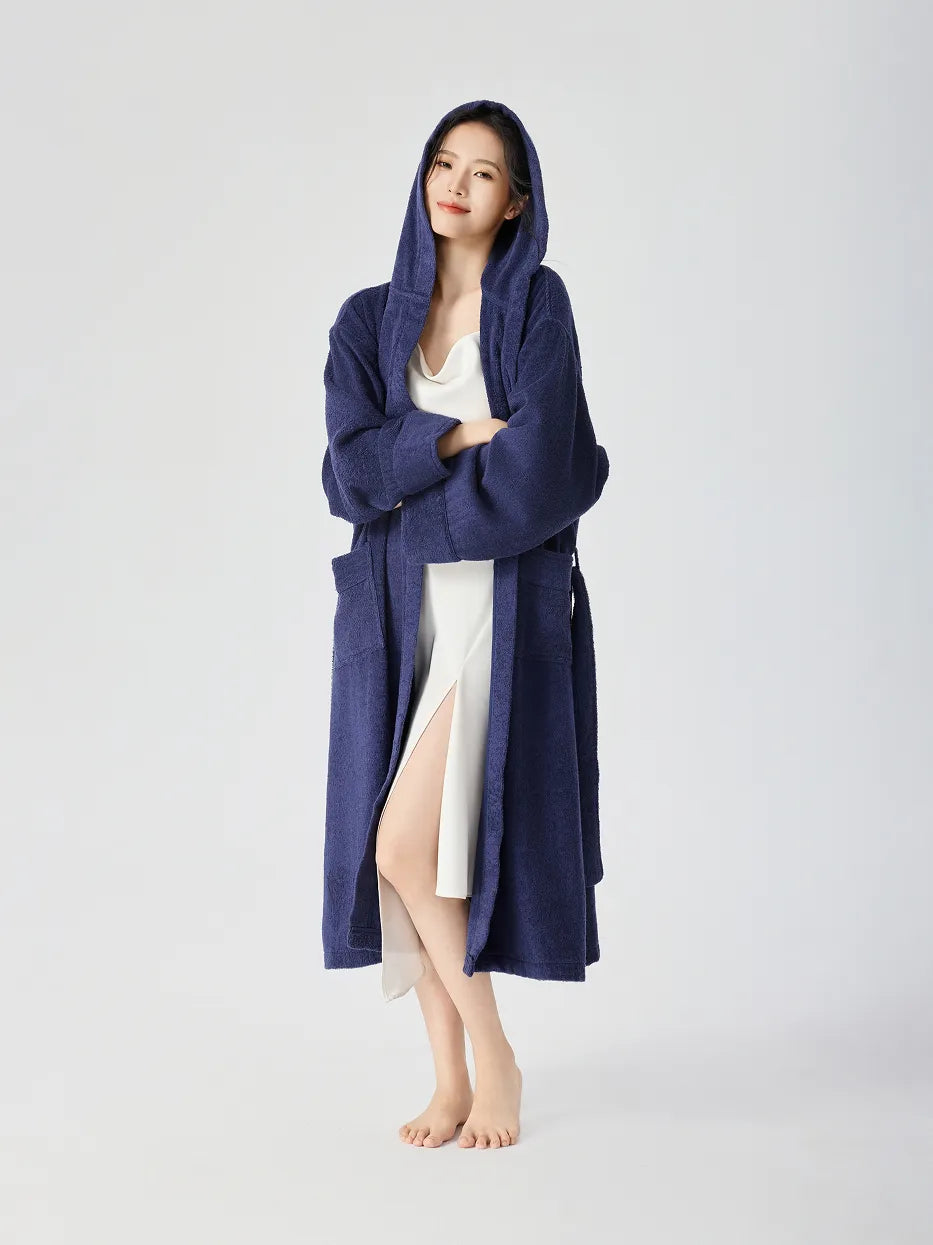 Barefoot woman wearing RESONG navy blue hooded terry cloth bathrobe with hood up, full length, arms crossed, over white slip dress with side slit, relaxed long fit, white studio background
