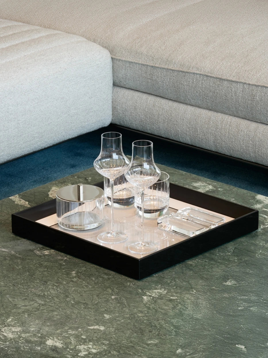 Italian minimalist reception layout shot of large black square tray with fluted crystal wine goblets & glass storage jar on green marble coffee table