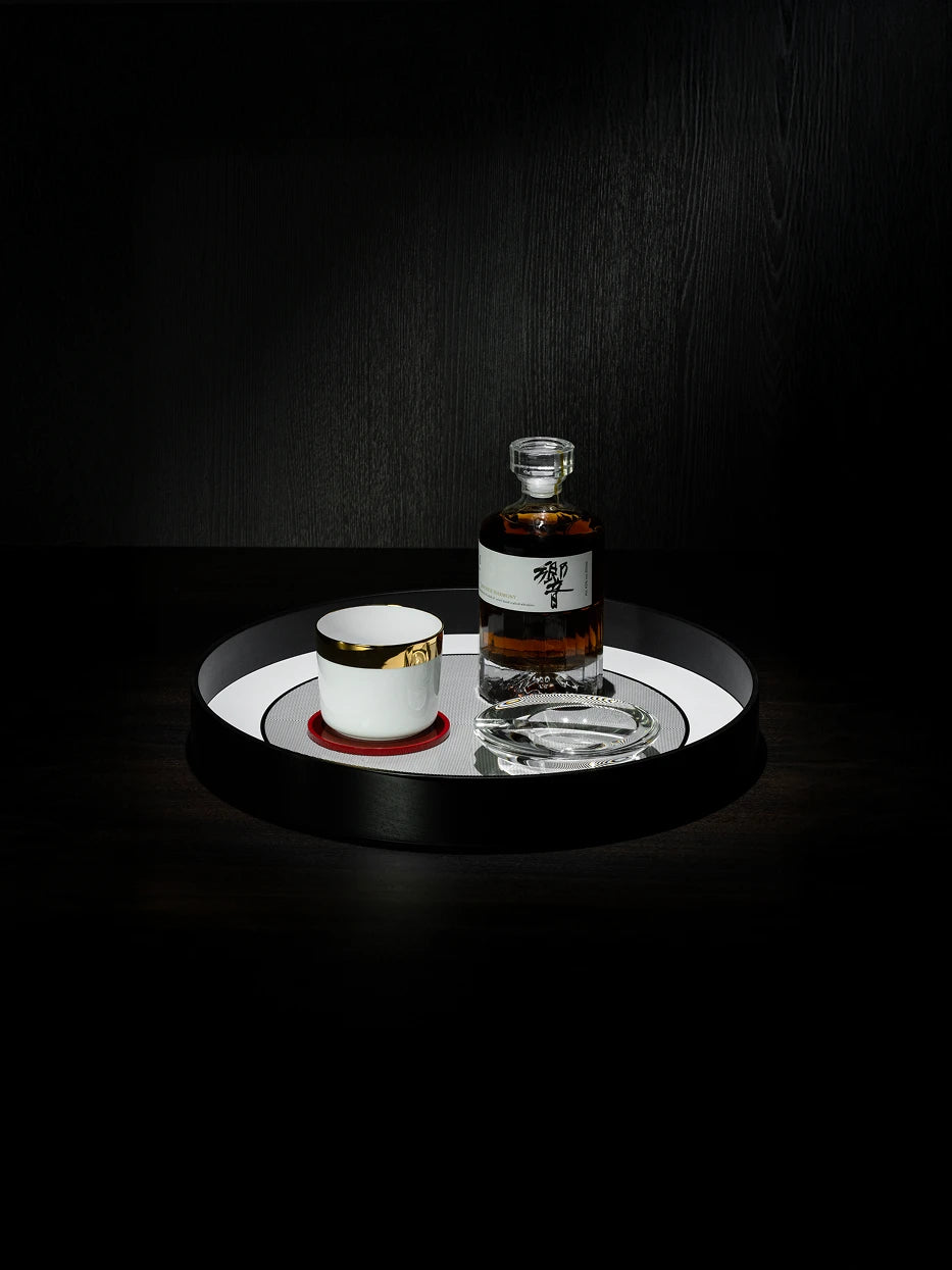 Dark tone men's wine layout shot of large black round tray with whiskey bottle, gold-rimmed mug & red coaster on dark wood grain table