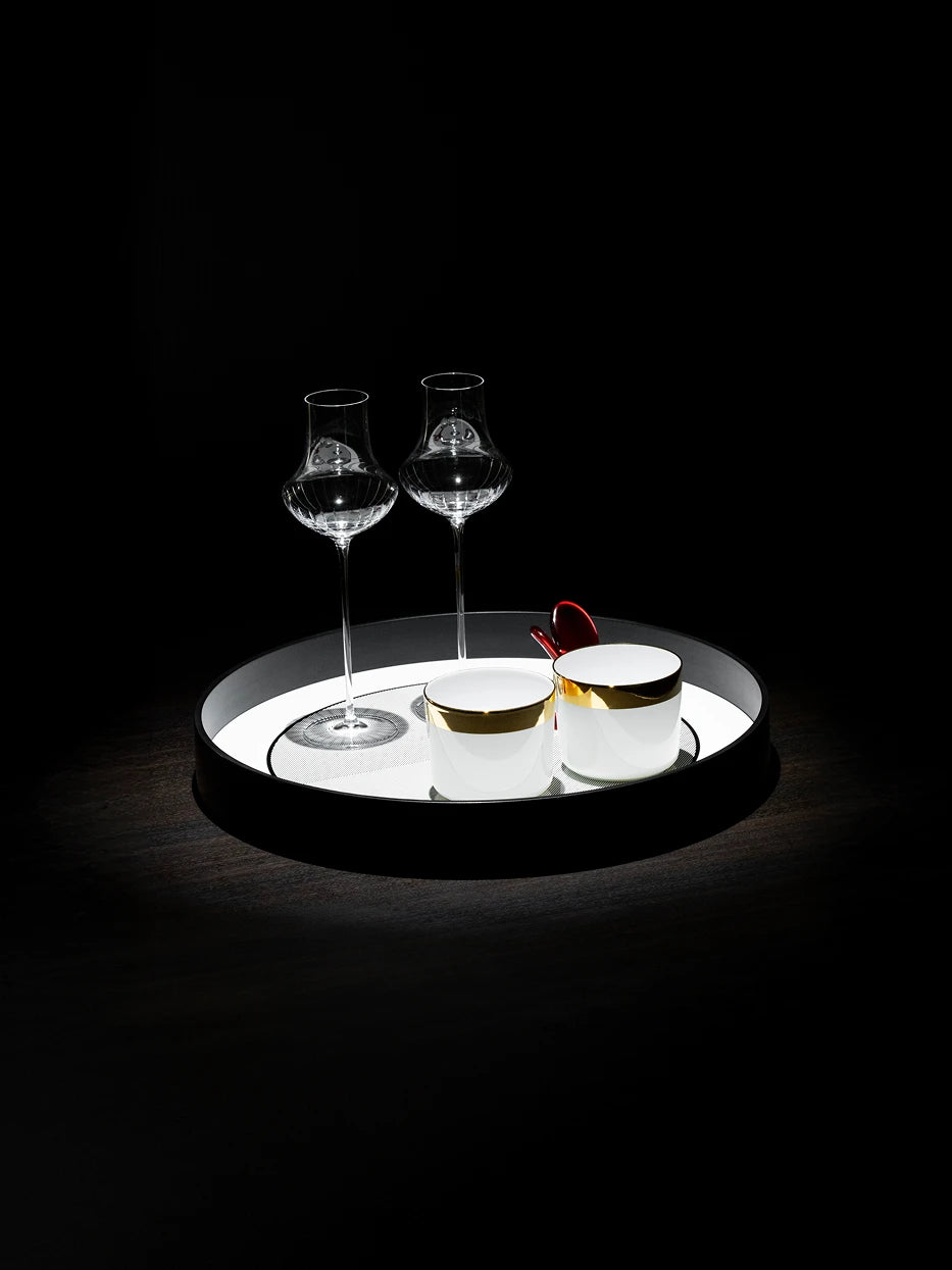 Dark tone texture still life shot of black round tray with fluted crystal wine goblets and gold-rimmed white mugs on dark solid wood table