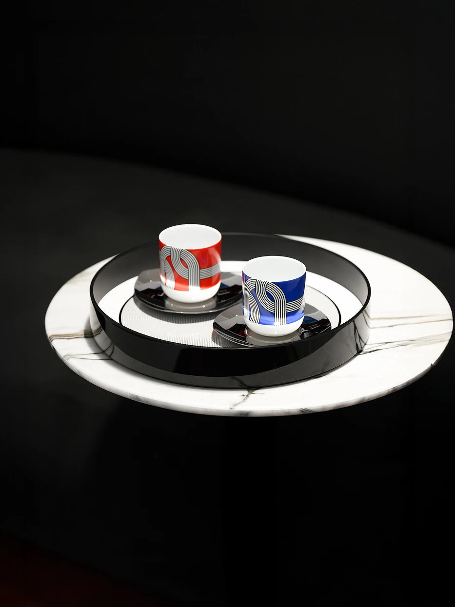 Modern minimalist reception shot of black medium round tray holding red & blue geometric printed mugs with matching coasters on black-white marble round coffee table