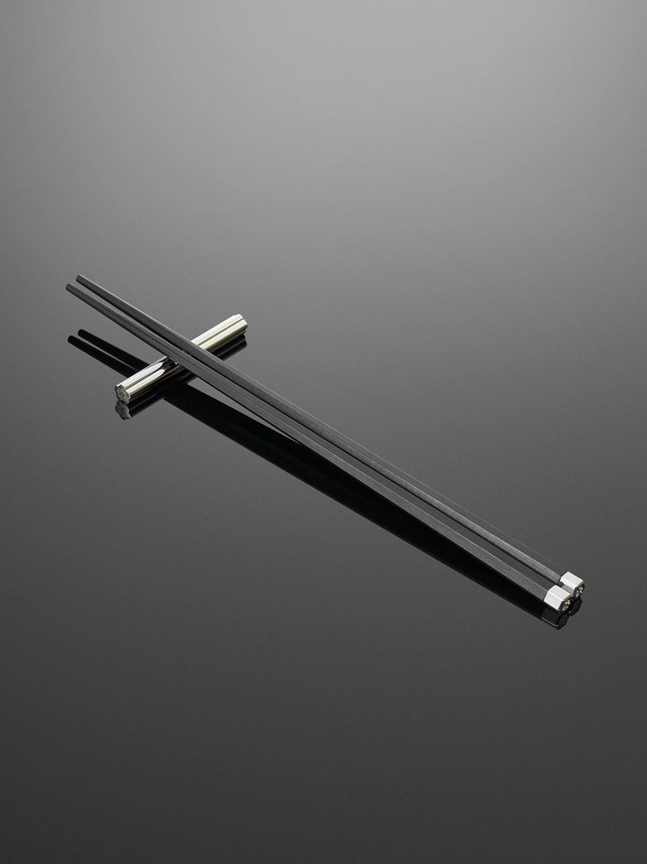 Minimal dark studio still life of single pair PAVILION ebony chopsticks laid on stainless steel rest, soft reflection on dark grey glossy table highlighting matte wood and embossed silver tip