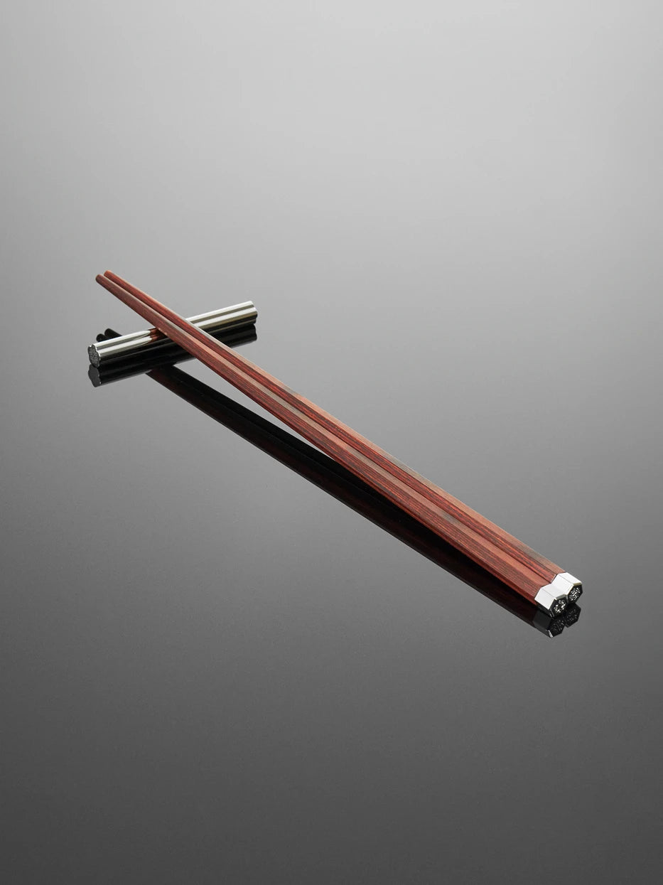 Minimal dark studio still life of single pair PAVILION rosewood chopsticks with stainless steel rest, soft reflection on matte grey table highlighting wood grain and alloy tip gloss