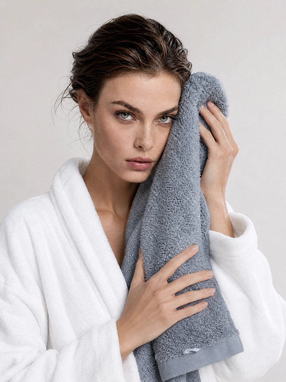 Half-body scene shot of model wearing white bathrobe holding gray cotton bath towel against cheek under plain minimalist background, shows soft skin-friendly touch, presents daily facial cleansing usage scene, intuitively displays delicate fluffy terry texture