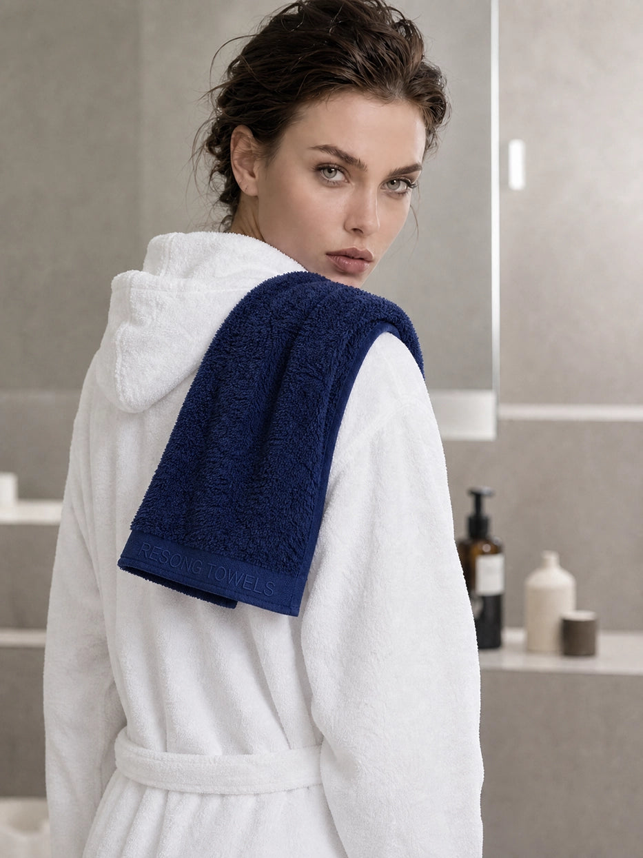 Scene shot of model wearing white bathrobe with R Collection navy blue cotton face towel draped over shoulder in minimalist stone bathroom, shows post-bath hair drying & facial cleansing usage scene, clear embroidered band detail, rich layered matching effect with contrast color bathrobe