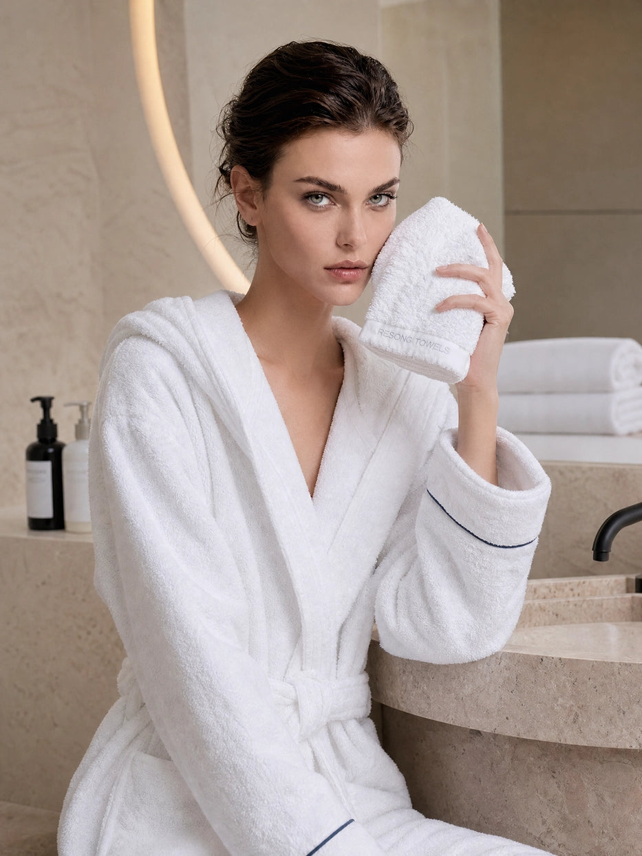 Scene shot of model wearing bathrobe holding R Collection white cotton face towel in minimalist stone bathroom, shows daily facial cleansing usage scene, clear embroidered band detail, matching set furnishing effect with same series bathrobe
