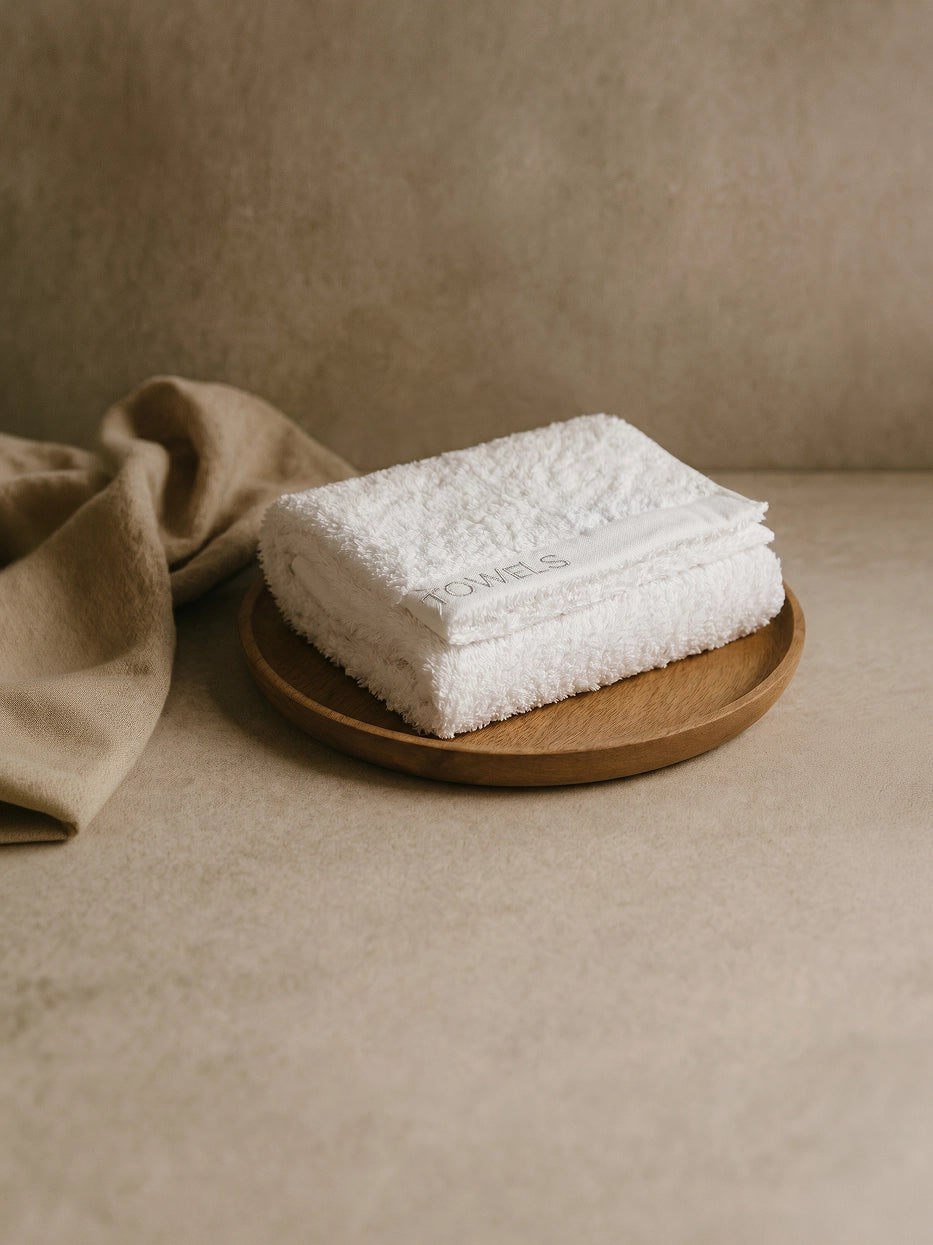 Wabi-sabi still life shot on beige micro-cement countertop with wooden tray, folded white R Collection cotton face towel matched with linen fabric, soft natural light creates minimalist bathroom atmosphere, shows interior display matching effect