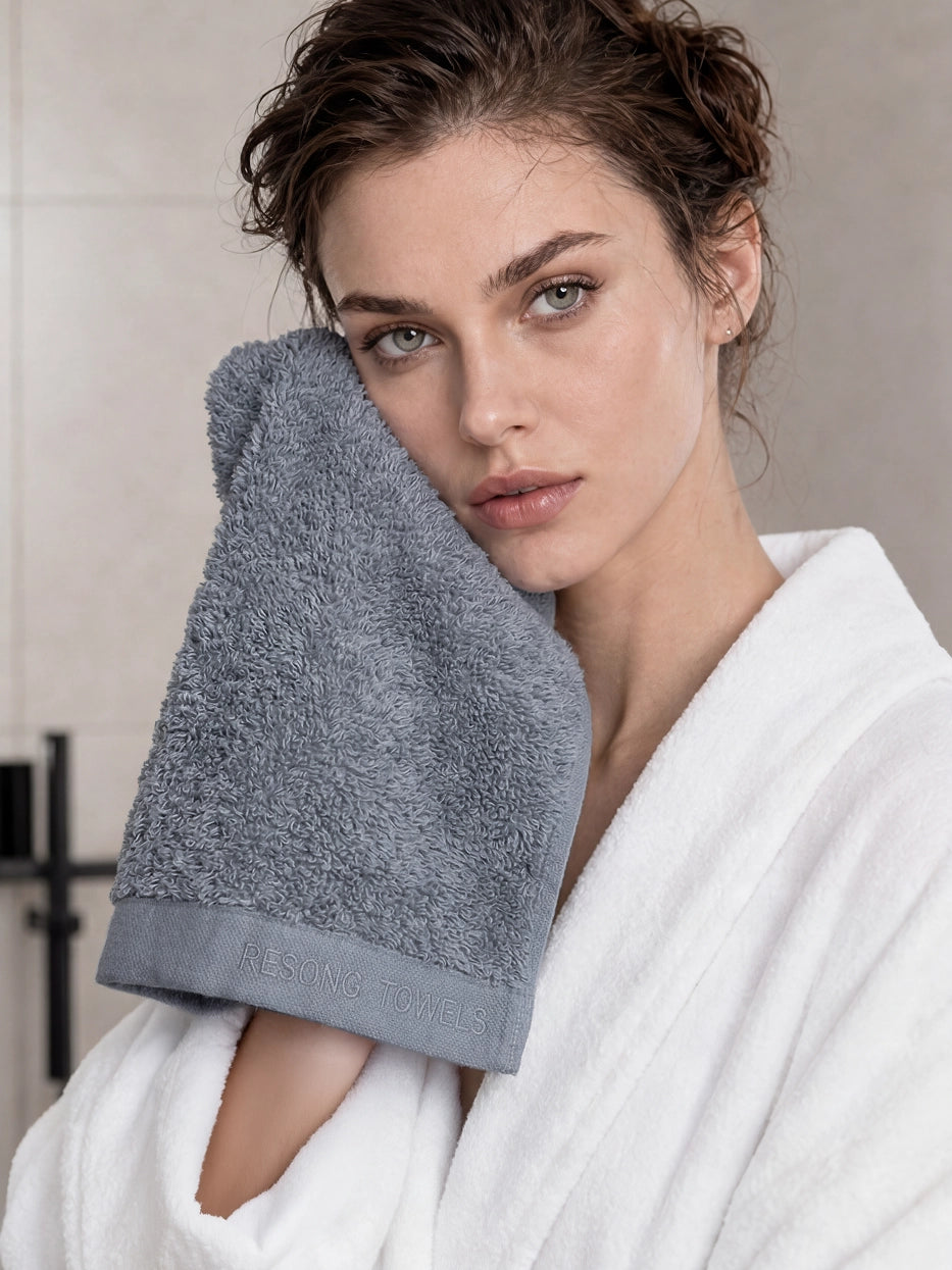 Half-body bathroom scene shot of model in white bathrobe holding grey face towel against cheek in light marble luxury bathroom, clear RESONG TOWELS embroidery on hem, shows multi-scene usage for facial cleansing & hair drying, highlights thick fluffy skin-friendly terry texture
