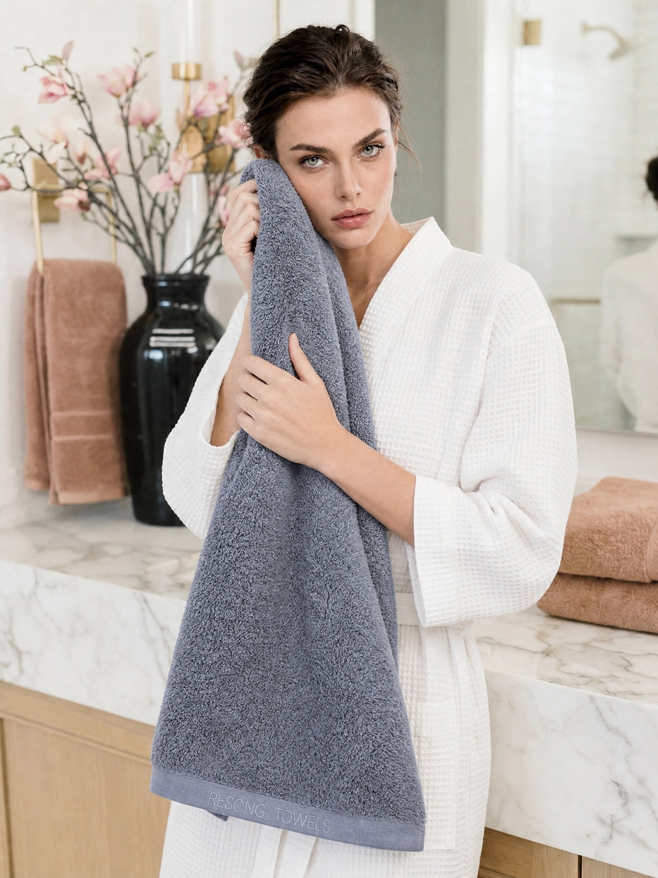 Half-body detail scene shot of model holding oversized grey bath towel in light marble luxury bathroom, fully reveals embroidered brand detail on hem, highlights 1400×700mm full wrap size & 640g thick fluffy terry, shows multi-scene post-shower drying usage, premium contrast matching between grey towel and white bathrobe