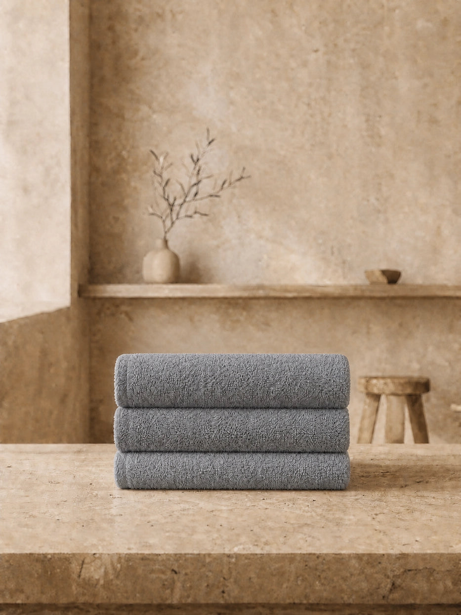 Wabi-sabi still life shot of three vertically stacked grey face towels on beige micro-cement countertop, unified solid grey tone soft and calm, matched with dried branch pottery decor to create minimalist bathroom vibe, shows full gift set display matching effect