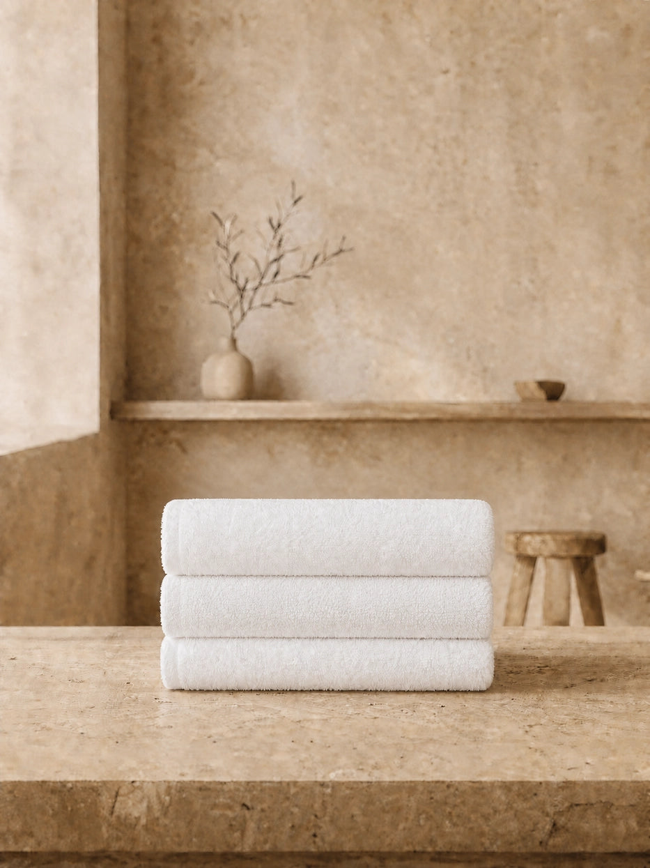 Wabi-sabi still life shot of three vertically stacked white face towels on beige micro-cement countertop, unified pure white tone clean and soft, matched with dried branch pottery decor to create healing minimalist bathroom vibe, shows full gift set display matching effect