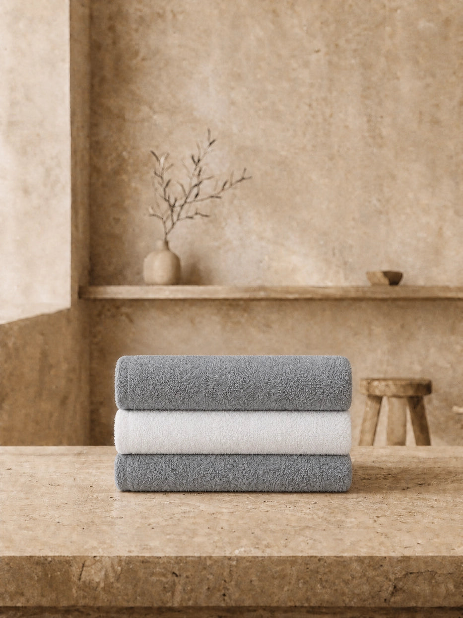 Wabi-sabi still life shot of three vertically stacked face towels on beige micro-cement countertop, staggered layered grey & white towels matched with dried branch pottery decor, creates soft neutral bathroom vibe, shows full gift set display matching effect