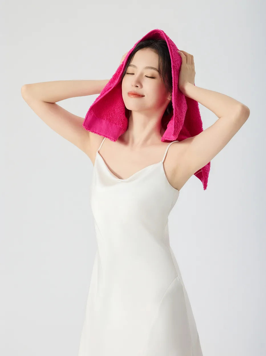 Woman in white slip dress, eyes closed draping RESONG rose red cotton terry face towel over head like a hood, soft fluffy face towel on model, white studio background
