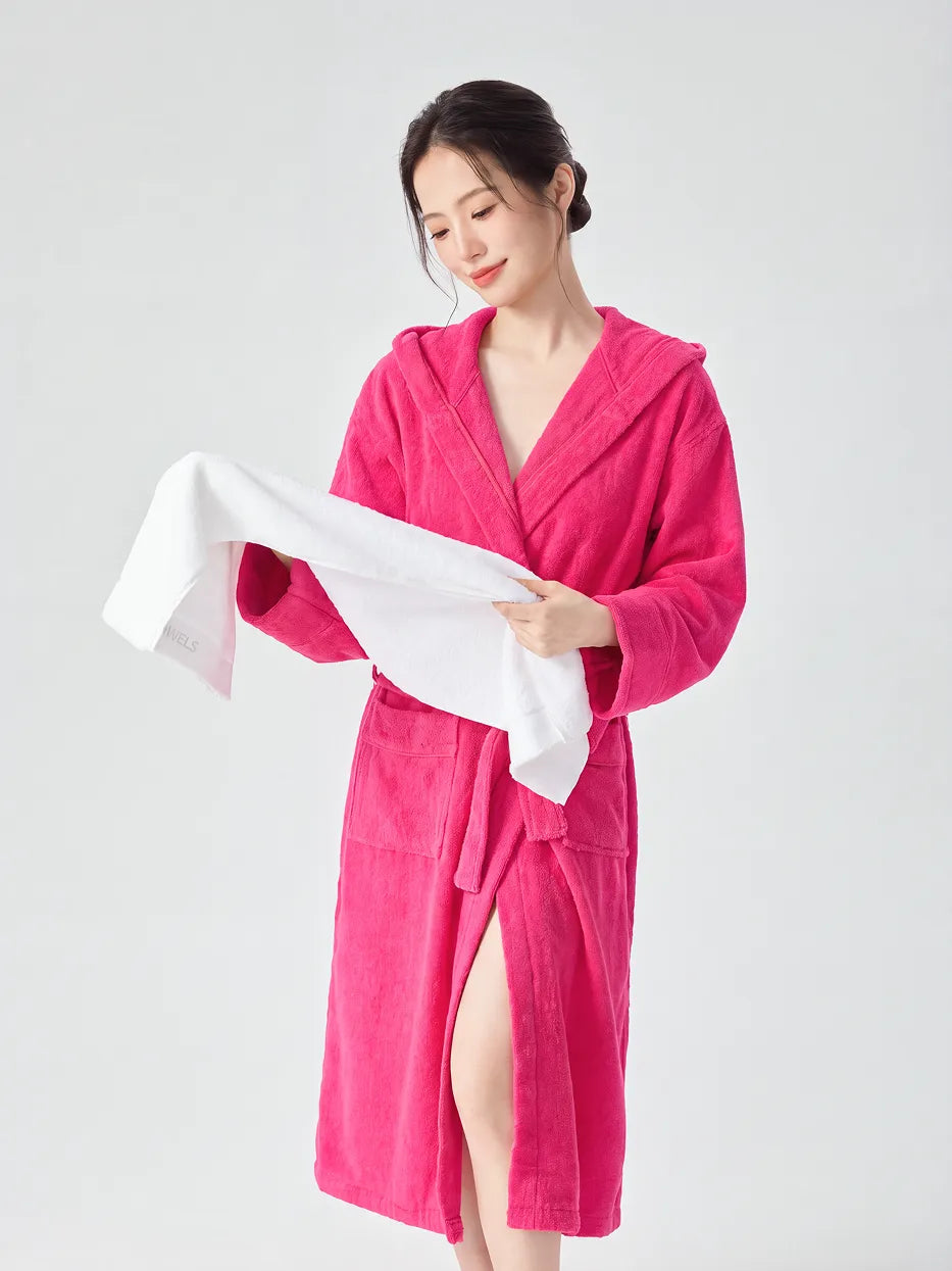 Woman wearing RESONG rose red hooded terry cloth bathrobe, unfolding a white cotton bath towel and looking down at its woven label, luxury bath towel and bathrobe gift set, white studio background
