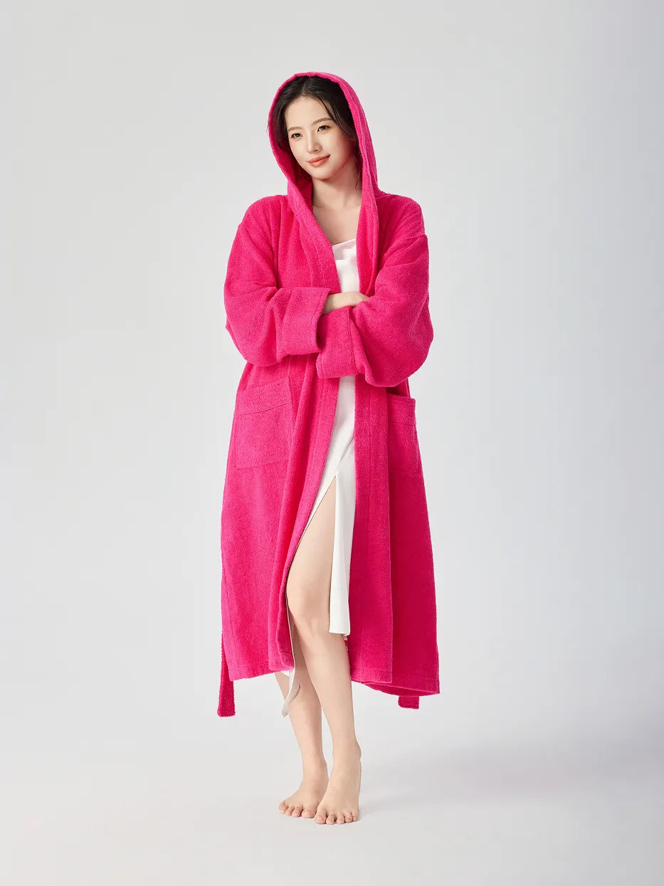 Barefoot woman wearing RESONG rose red hooded terry cloth bathrobe with hood up, arms crossed, full length, white slip dress underneath, relaxed long bathrobe with patch pockets, white studio background
